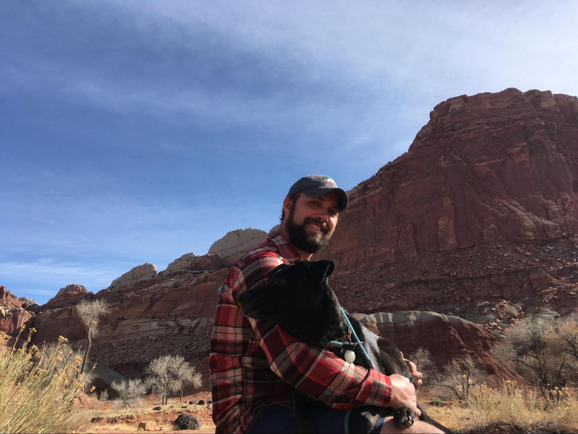 Andy sits outside with a dog on his lap in front of a large, rocky mountainous landscape under a clear blue sky. He wears a plaid shirt and a baseball cap, smiling at the camera. The dog looks content and is embraced warmly. Sparse vegetation surrounds them, showcasing his love for outdoor hobbies amidst nature’s beauty.
