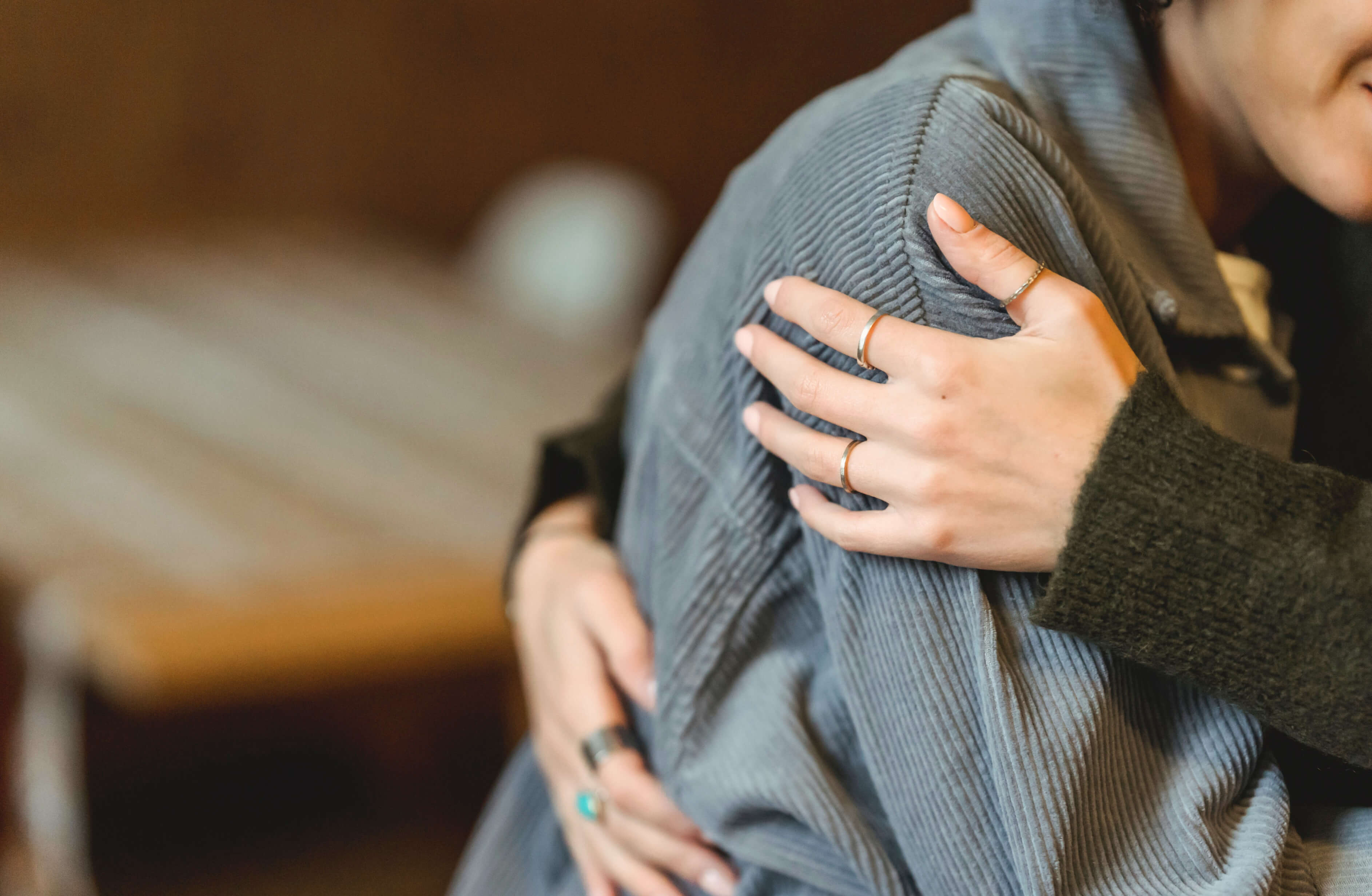 A close-up of two people embracing. One person is wearing a gray jacket, and the other has their arms wrapped around them, showing hands with several rings. The background appears to be indoors with a blurred, warm ambiance, evoking a moment of support that could signify comfort during California bereavement leave.