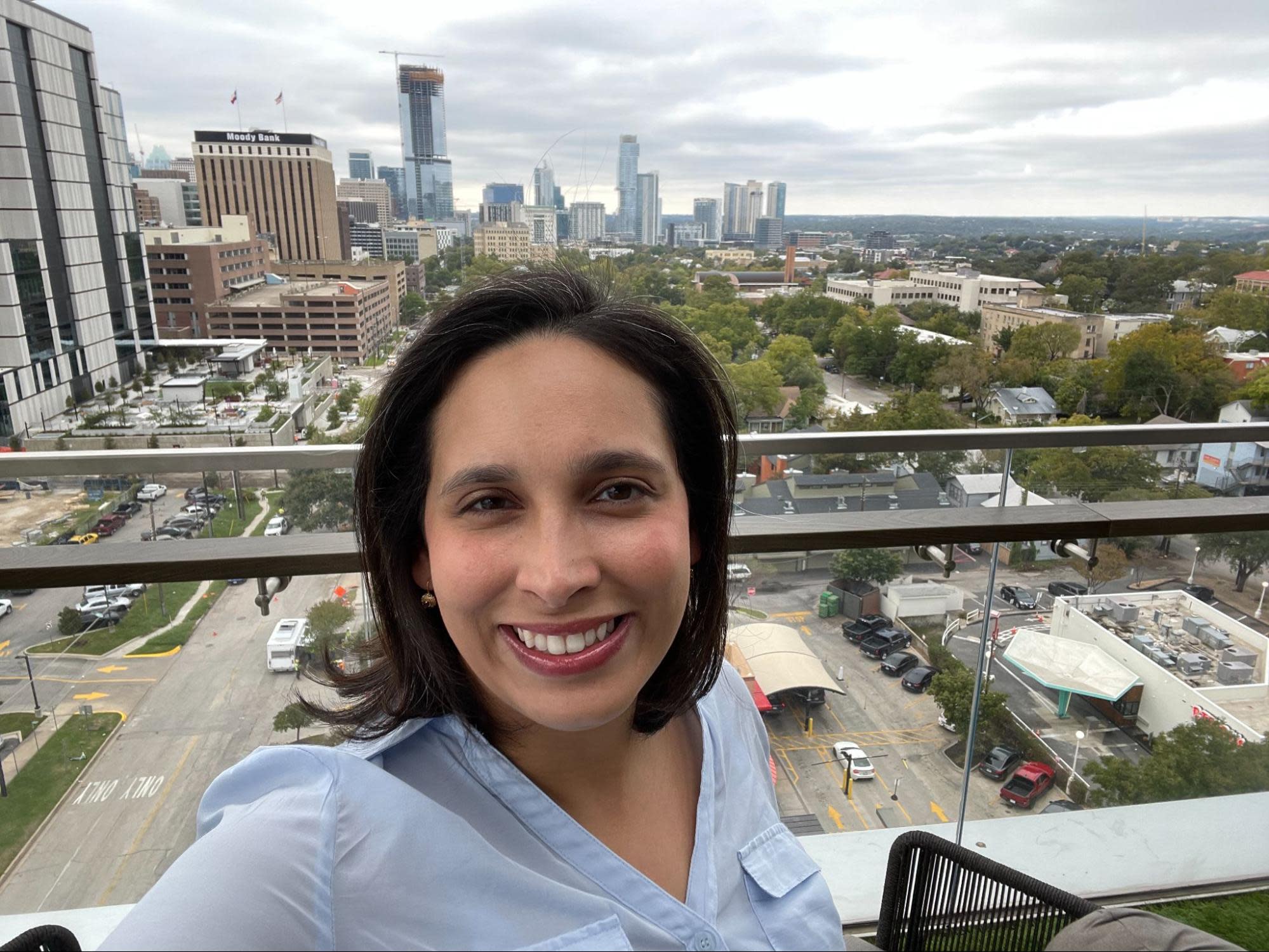 A person with shoulder-length dark hair and a light blue shirt is taking a selfie on a rooftop. The background shows a cityscape with various buildings, trees, and cars on the streets below, under a cloudy sky. Their passion for tropical gardening is evident in the lush plants nearby.
