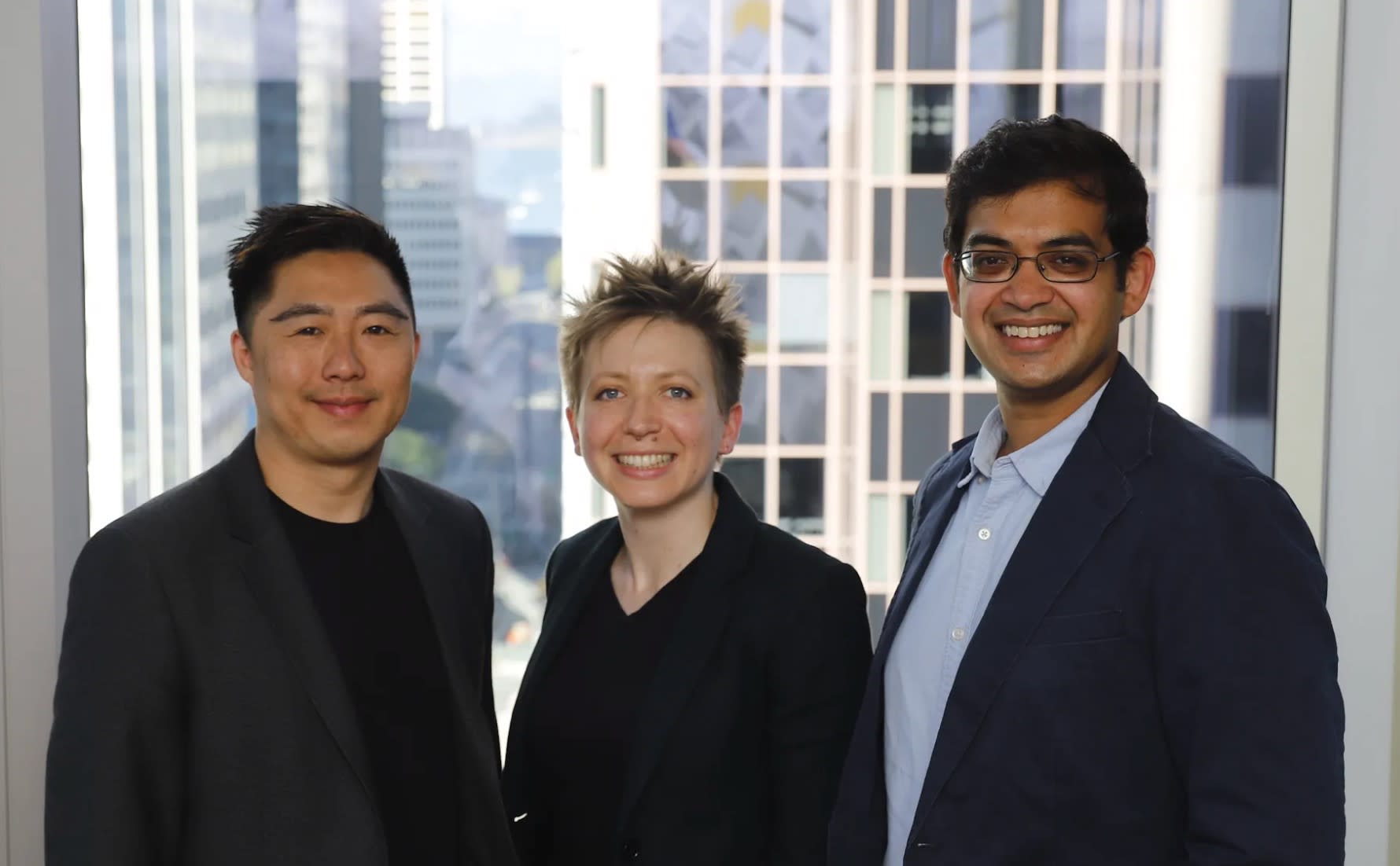 Three people are standing together in front of a window overlooking an urban cityscape with tall buildings. They are all smiling and wearing business casual attire. The person on the left is wearing a dark blazer, the person in the middle a black shirt, and the person on the right a blue blazer, discussing their recent Sparrow funding success.